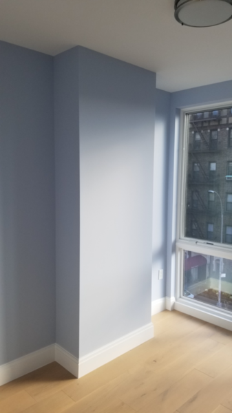 Empty room corner with pale blue gray painted walls, white baseboards, and a large window facing brick buildings