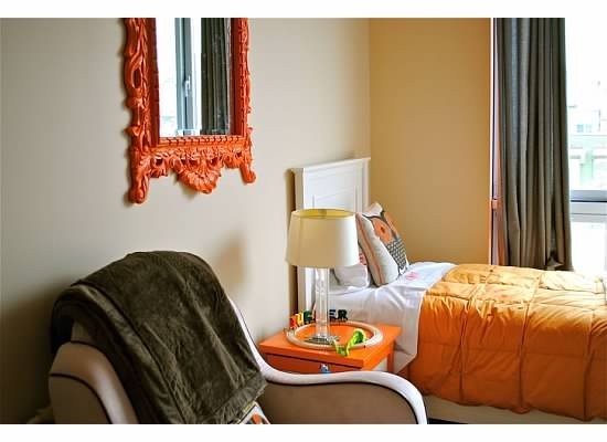 Bedroom corner with cream walls, orange bedding, an orange side table with lamp, and a carved orange mirror